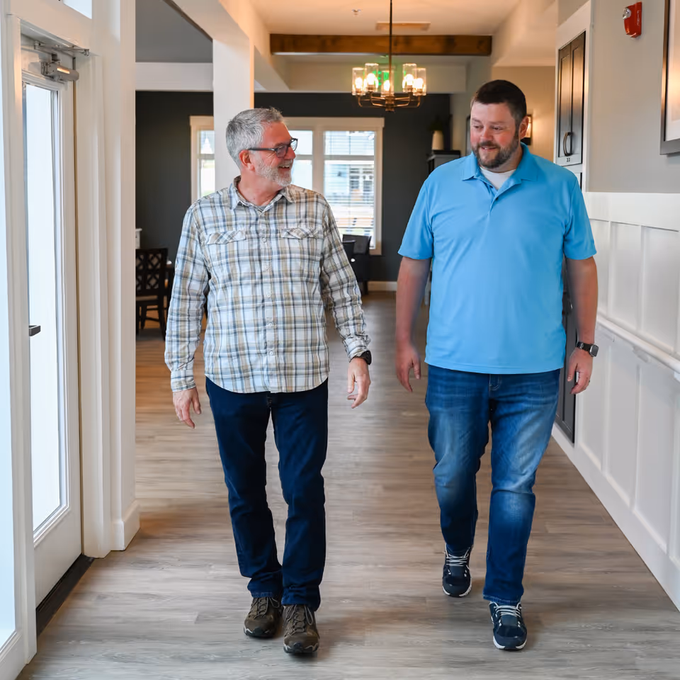 Two men walking and talking in a well-lit hallway with wooden flooring and white walls. One man is wearing a plaid shirt and glasses, and the other is wearing a light blue polo shirt. There is a chandelier hanging from the ceiling and a dining area visible in the background.