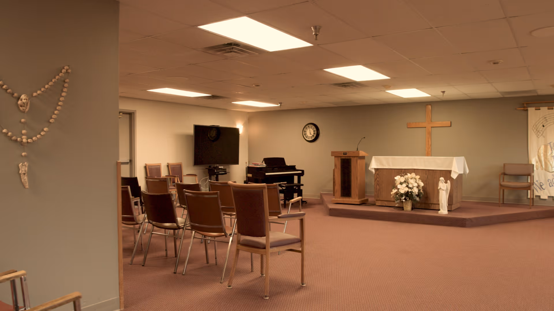 Interior view of a small chapel or worship room with rows of chairs facing a raised platform. The platform has a wooden podium, an altar covered with a white cloth, a wooden cross on the wall, a bouquet of white flowers, and a small statue. There is a piano and a wall clock in the background, and a decorative rosary hanging on the left wall.