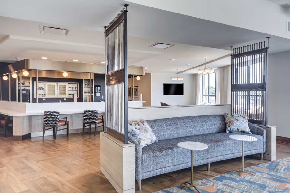 Open communal lounge with a gray sofa, two small round tables, a bar counter with stools, pendant lights, and a wall-mounted TV.