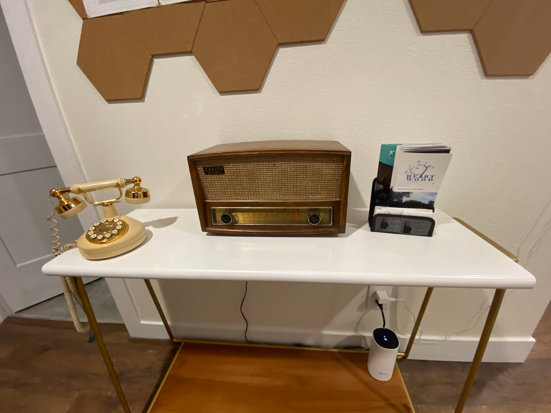 A white table with gold legs against a white wall with brown hexagonal wall decorations. On the table, there is a vintage rotary telephone, a vintage Zenith AM/FM radio, and a black holder containing brochures and cards. Below the table, a white cylindrical device is plugged into a wall outlet.
