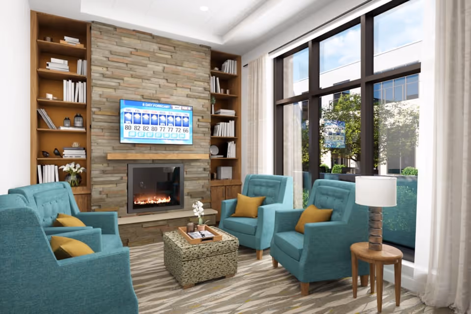 A cozy living room with four teal armchairs each with a mustard yellow pillow, arranged around a patterned ottoman. A stone fireplace with a mounted flat-screen TV displaying a 6-day weather forecast is centered on the wall, flanked by built-in wooden bookshelves filled with books and decorative items. Large windows with sheer curtains let in natural light and show greenery outside. A small wooden side table with a lamp is next to one of the armchairs.