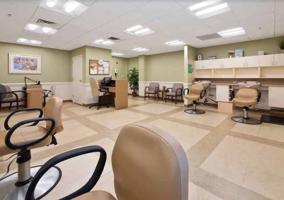 Interior view of a senior living facility's salon area with beige salon chairs, a reception desk with a computer, a few waiting chairs along the wall, green walls with white trim, and overhead fluorescent lighting.