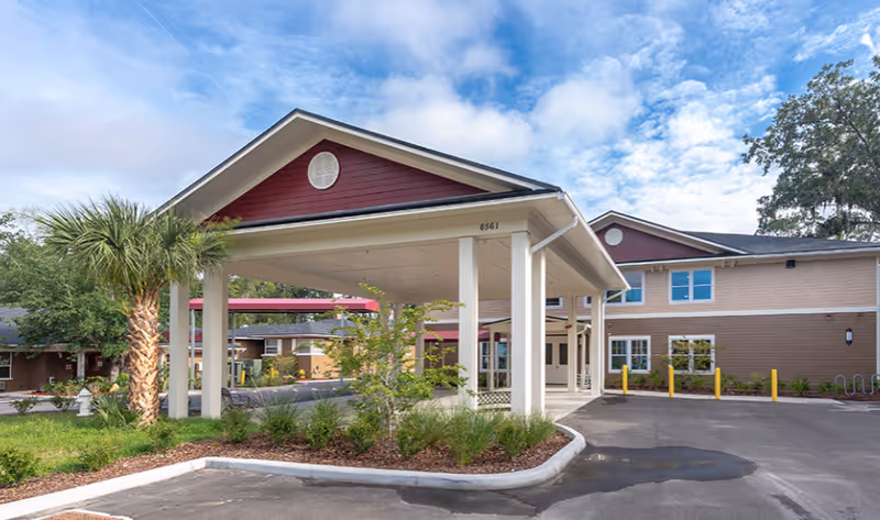 Exterior view of a senior living facility with a covered entrance supported by white columns. The building has a two-tone brown and beige facade with red gable accents and multiple windows. There is a palm tree and landscaped greenery near the entrance, and a paved driveway in front.