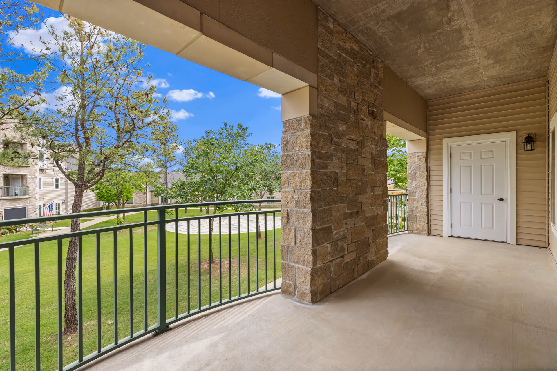 Covered second-floor balcony with stone columns and green metal railing overlooking a grassy courtyard and nearby buildings.