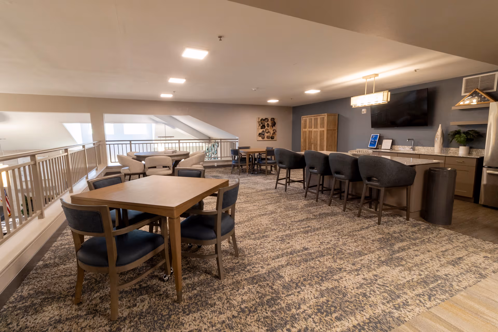 A spacious interior common area with multiple seating arrangements including tables with chairs and a kitchen island with bar stools. The room features a carpeted floor, a large flat-screen TV mounted on a dark accent wall, and modern lighting fixtures. There is a railing overlooking a lower level on the left side.