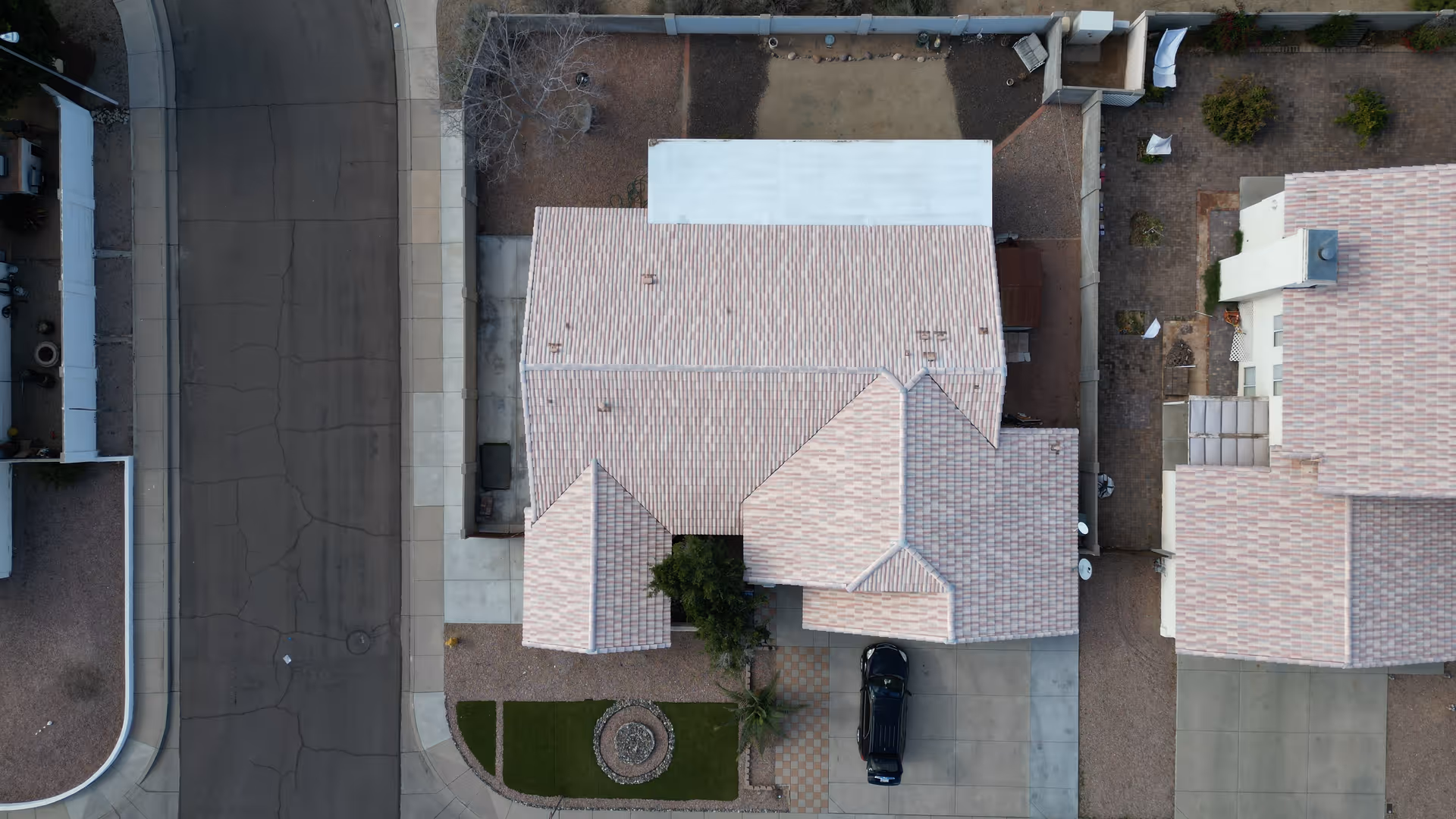 Aerial view of a residential building with a tiled roof, driveway with a parked black car, small landscaped front yard with a circular stone feature, and surrounding sidewalks and streets.
