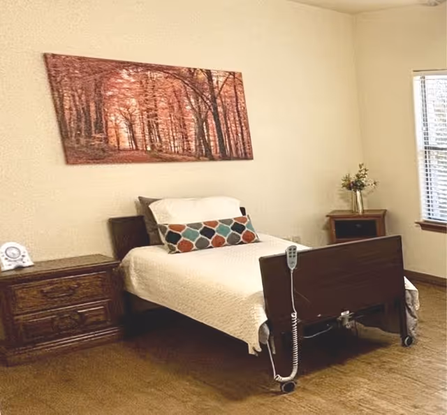 A simple bedroom with a single hospital-style bed featuring a white quilt and a colorful geometric pillow. The bed has a remote control attached to the footboard. To the left of the bed is a wooden nightstand with a clock on it, and to the right is a small wooden table with a vase of flowers. A large framed picture of a forest with autumn-colored trees hangs on the wall above the bed. A window with blinds is on the right side of the room, letting in natural light.