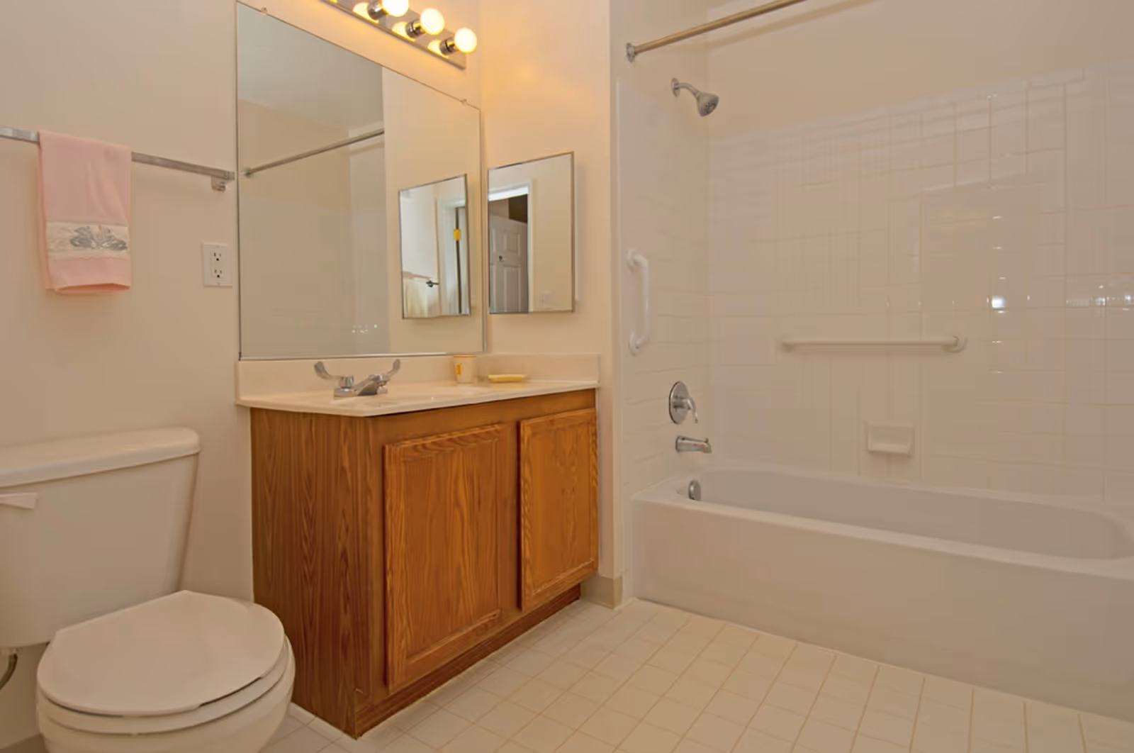A clean bathroom featuring a white toilet, a wooden vanity with a sink, a large mirror above the sink, a bathtub with white tiled walls, a showerhead, and grab bars for safety. A pink towel hangs on a towel rack next to the toilet.
