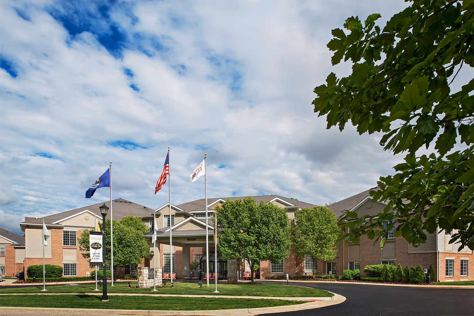 Exterior view of American House Park Place senior living facility with a circular driveway, three flagpoles displaying flags, well-maintained green trees and shrubs, and a partly cloudy sky.