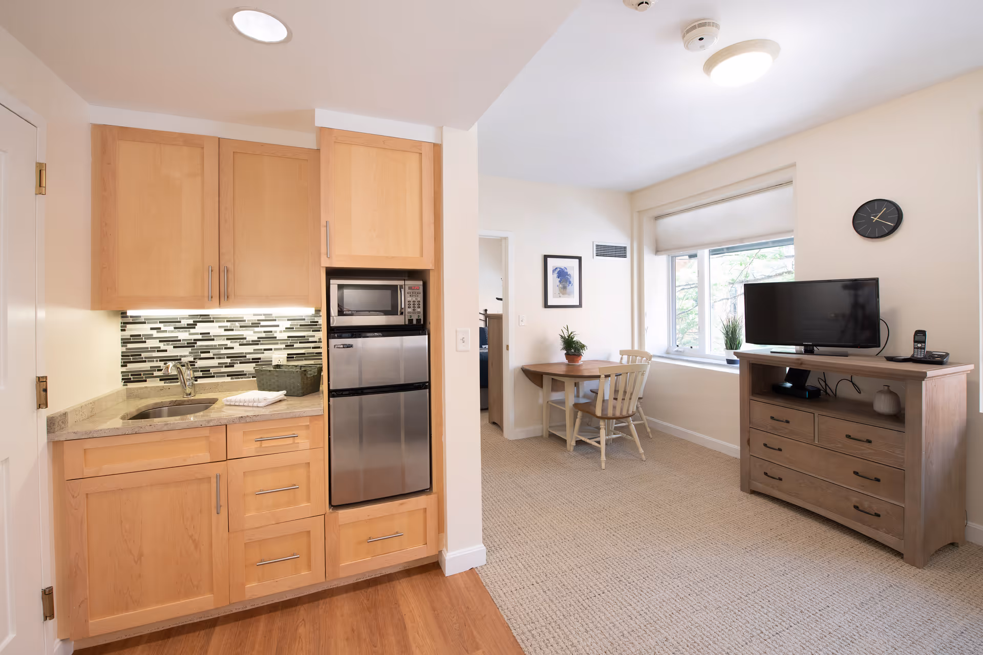 A bright and cozy assisted living apartment featuring a small kitchenette with wooden cabinets, a microwave, and a mini refrigerator. Adjacent to the kitchenette is a dining area with a round wooden table and two chairs, a window with a roller shade, and a wooden dresser with a flat-screen TV and a cordless phone on top. The walls are painted white, and the floor transitions from wood in the kitchenette to carpet in the living area.