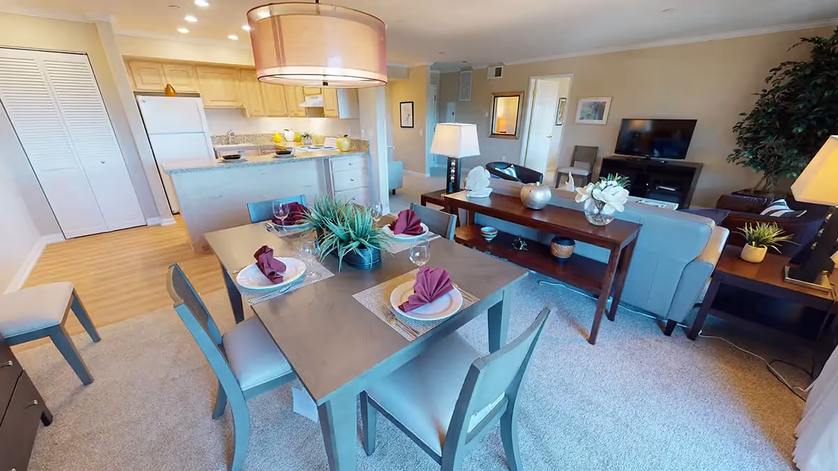 A bright and spacious open-concept living and dining area in a senior living facility. The dining table is set with four place settings, each with a plate, glass, and folded maroon napkin. A modern light fixture hangs above the table. Behind the dining area is a kitchen with light wood cabinets, a refrigerator, and a countertop with decorative items. The living room features a sofa, armchairs, a TV on a stand, a console table with decorative objects, and a large potted plant in the corner. The room has beige walls and carpeted floors with some hardwood near the kitchen.