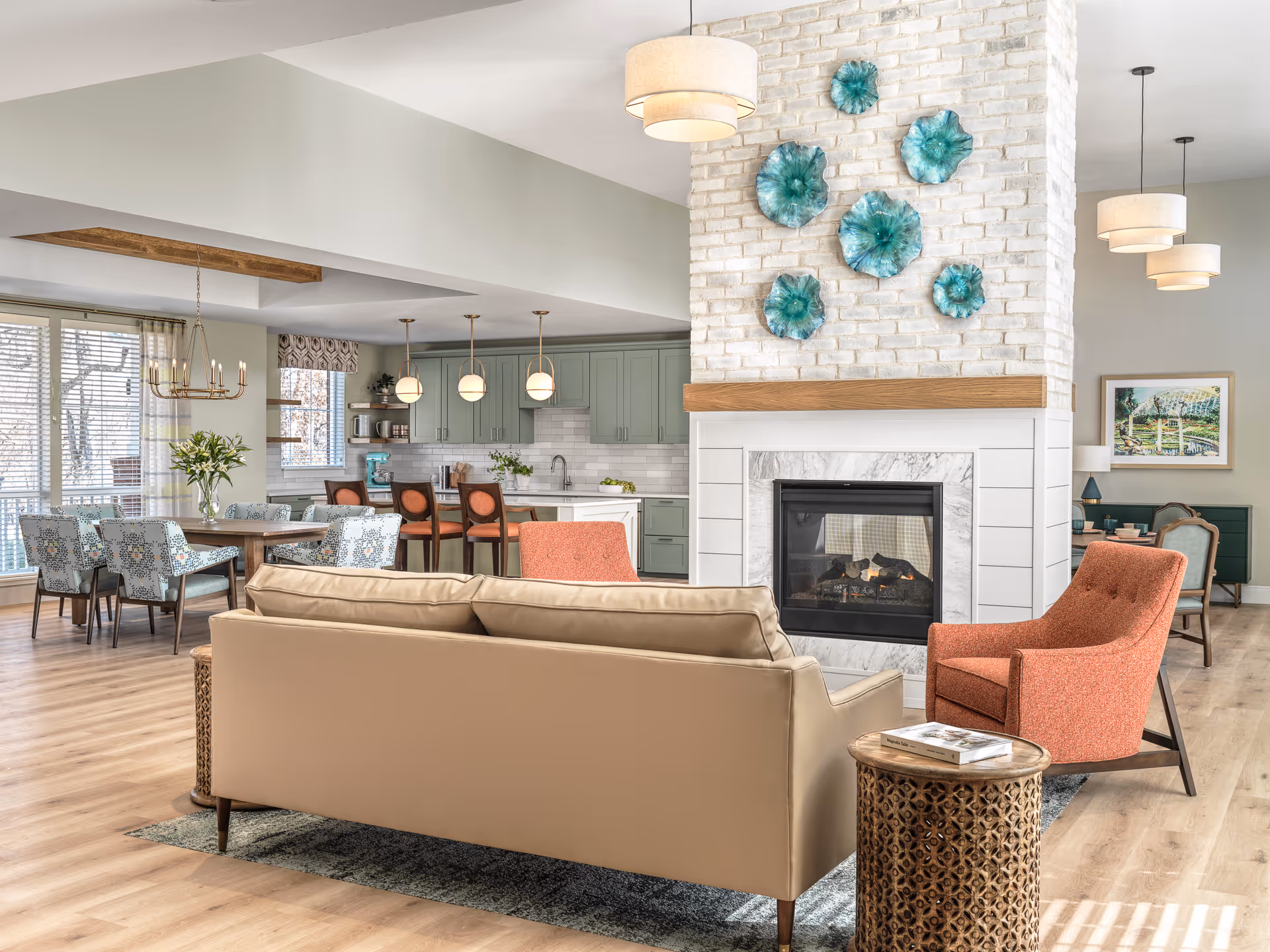 A bright and spacious living area in a senior living facility featuring a beige sofa and an orange armchair arranged around a modern fireplace with a white brick surround and decorative blue-green wall art above it. In the background, there is a dining area with a wooden table and patterned chairs, and a kitchen with light green cabinets, a white countertop, and pendant lights hanging above the island. The room has large windows with blinds and light wood flooring throughout.