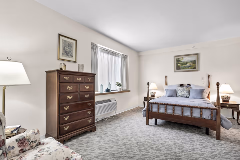 A bright bedroom with a wooden four-poster bed made with light blue bedding and pillows. On either side of the bed are matching wooden nightstands with table lamps. A wooden chest of drawers stands against the wall near a window with sheer curtains. A floral armchair and a floor lamp are partially visible in the foreground. The room has light-colored walls and carpeted flooring.