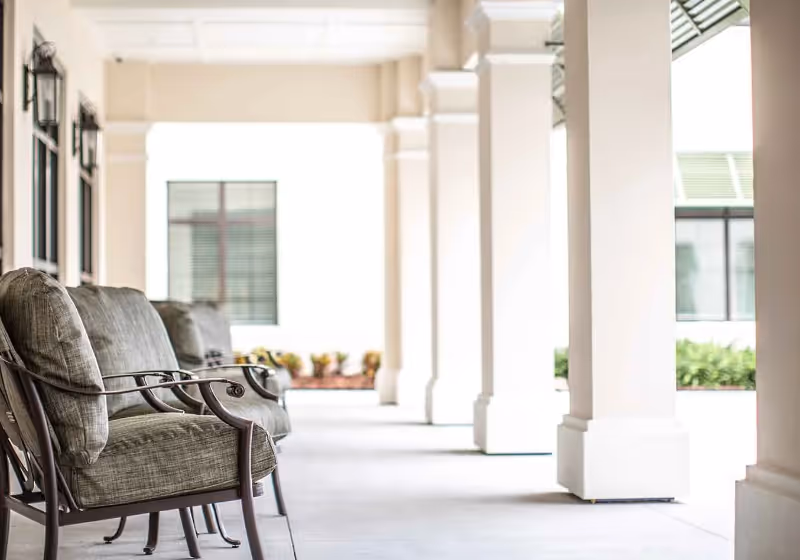 A covered outdoor patio area with cushioned metal chairs lined up along the left side, white columns supporting the roof, and windows with green shutters on the building wall in the background.