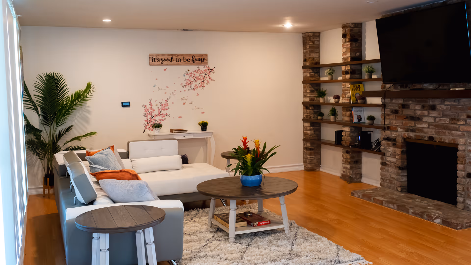 A cozy living room with a sectional sofa adorned with cushions and a throw blanket, a round wooden coffee table with a potted plant, and a side table. The room features a brick fireplace with built-in shelves holding decorative items and books. A wall decoration reads 'it's good to be home' above a small white console table. The floor is wooden, and there is a large green plant in the corner near a window with blinds.