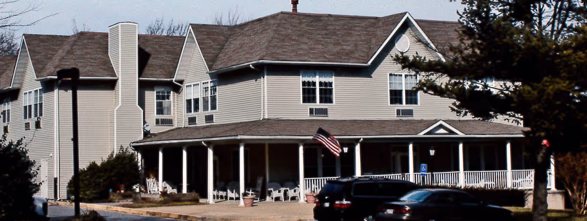 Front exterior of a two-story assisted living building with a wraparound porch, American flag and parked cars.
