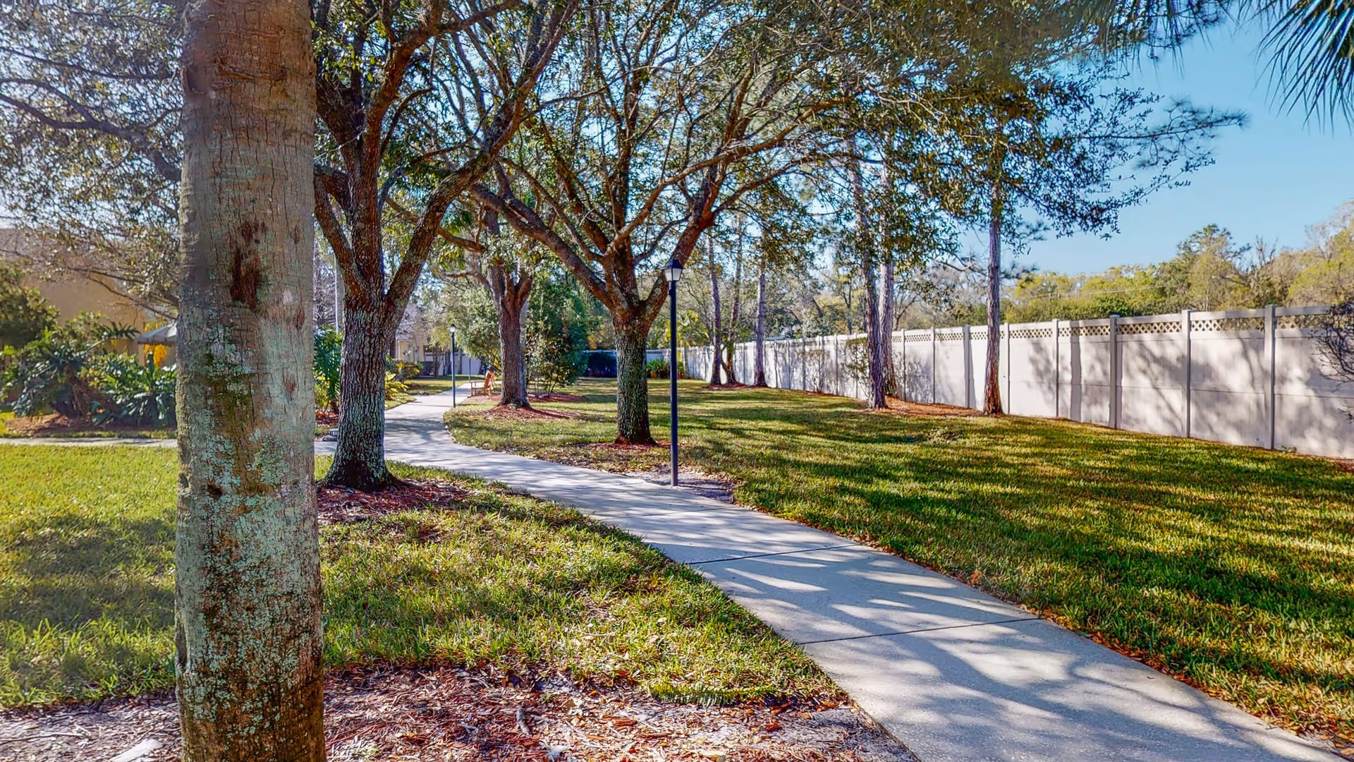 A paved walking path winding through a grassy outdoor area with several trees and lamp posts, bordered by a white fence on one side and landscaping on the other under a clear blue sky.