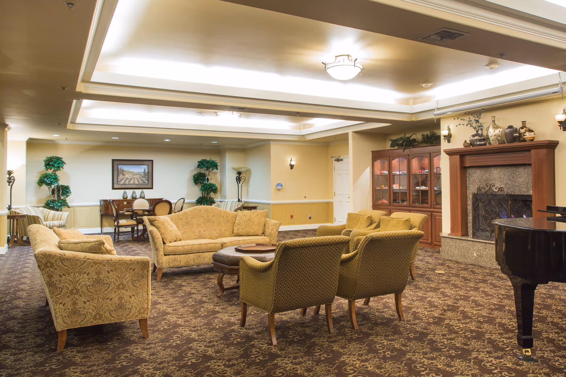 A spacious and elegantly furnished living room with patterned carpet, a set of upholstered sofas and armchairs arranged around a wooden coffee table, a fireplace with decorative items on the mantel, a glass-front cabinet, a grand piano, and a small dining table with chairs in the background. The room is softly lit with ceiling lights and wall sconces.