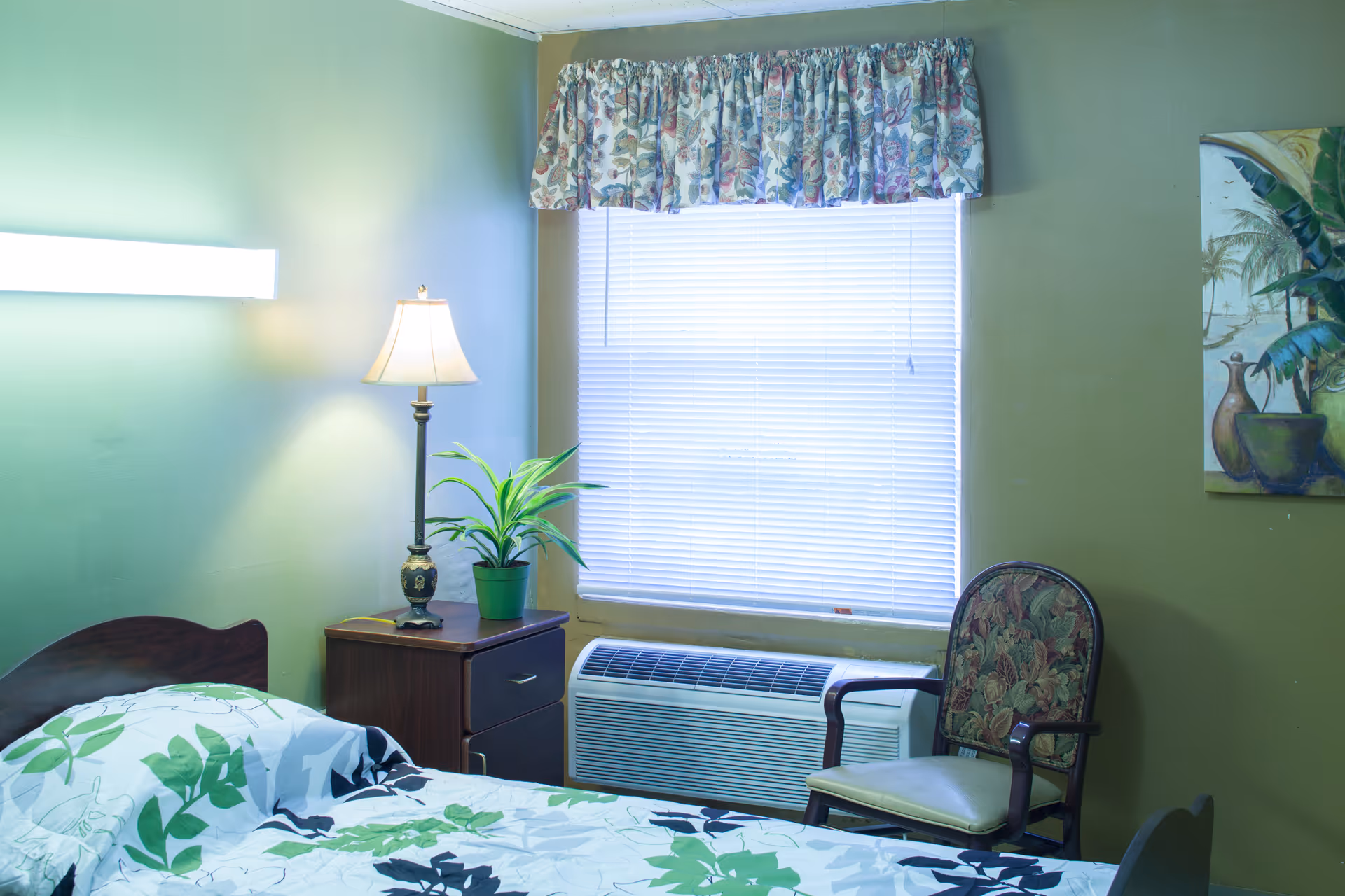 A small bedroom with a bed, bedside table and lamp, a potted plant, a chair, and a window with blinds above an air conditioner.