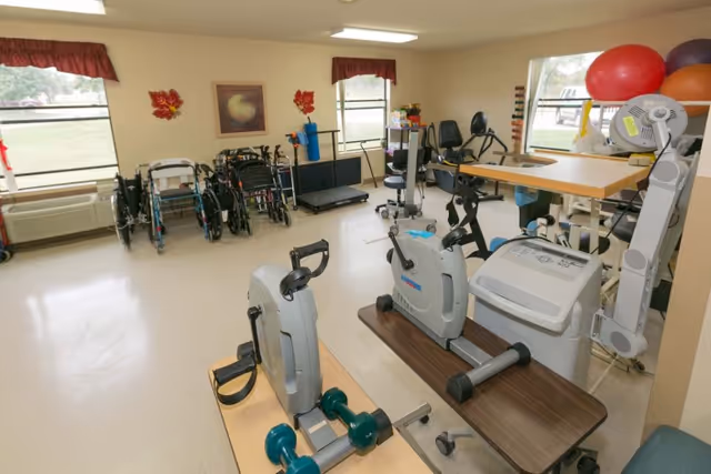 A rehabilitation or exercise room in a senior living facility with exercise bikes, wheelchairs, a treadmill, exercise balls, and other physical therapy equipment. The room has beige walls, two windows with red valances, and a light-colored floor.