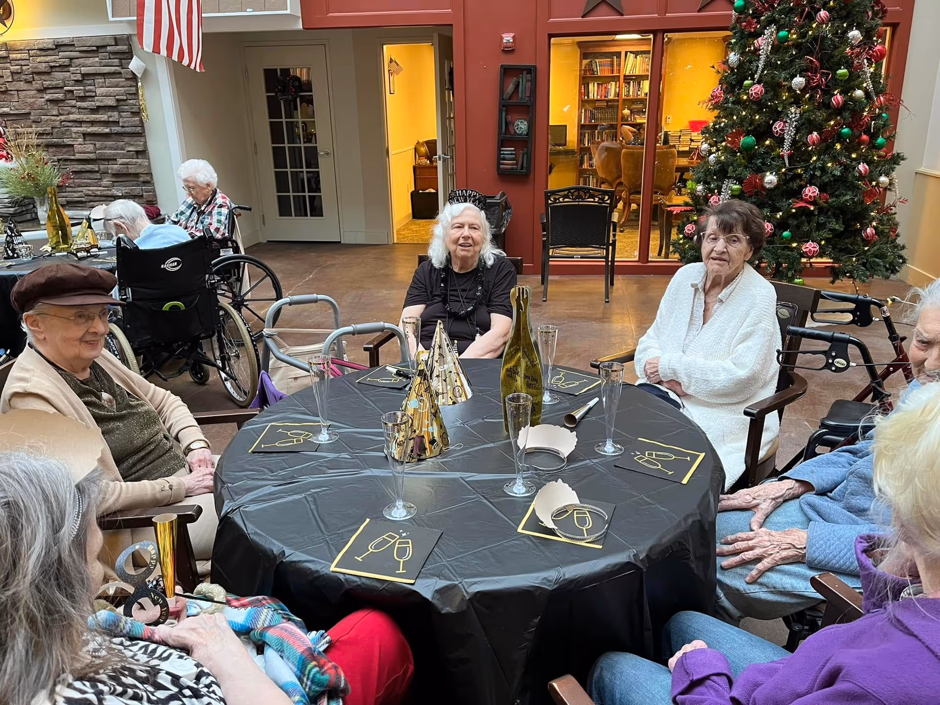 A group of elderly people sitting around a round table covered with a black tablecloth decorated with gold party hats, napkins with champagne glass designs, and empty champagne flutes. They are in a communal indoor area with a Christmas tree decorated with red and green ornaments in the background. Some individuals are in wheelchairs, and the setting appears festive and social.