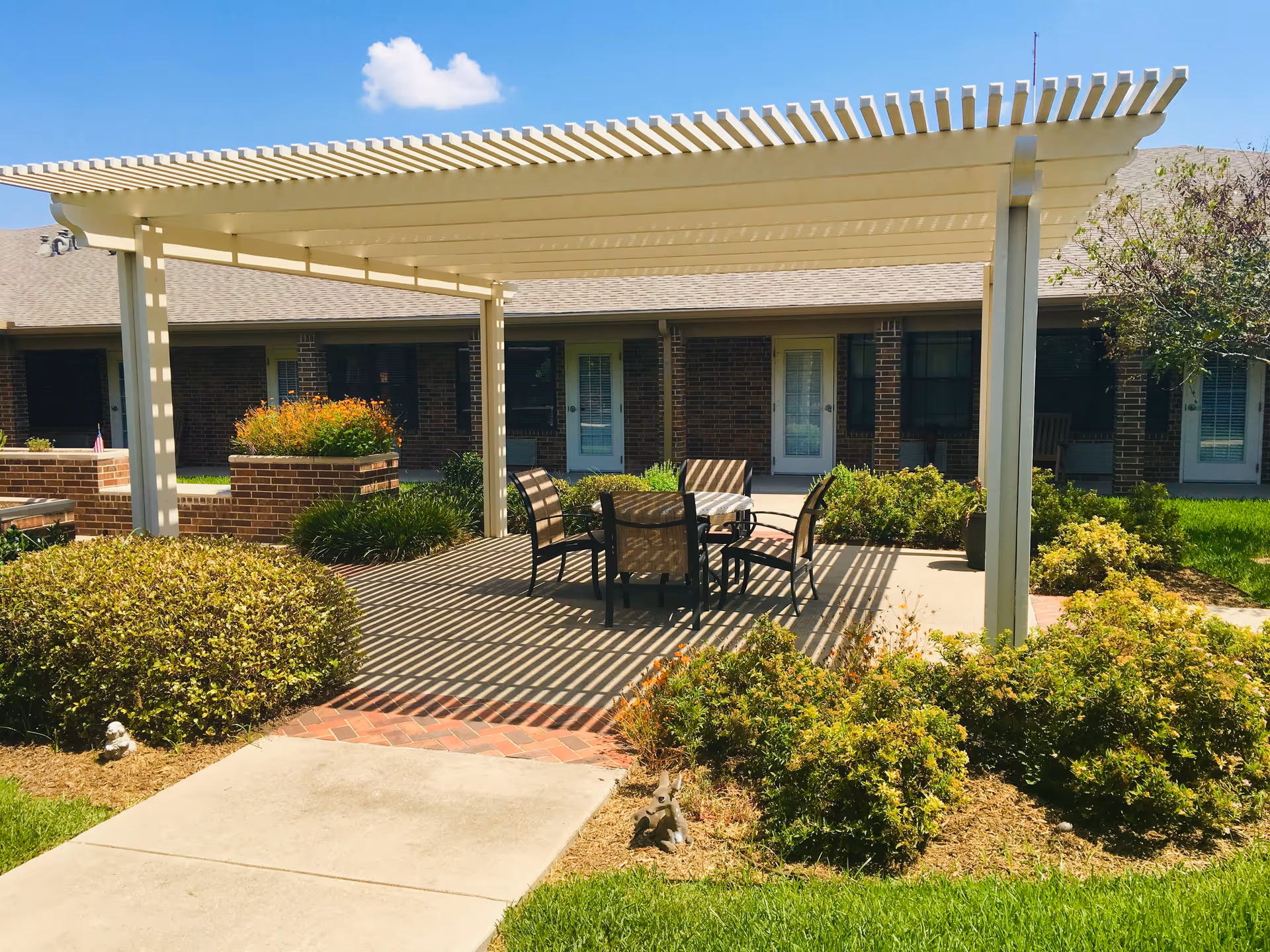 Outdoor patio area at Bella Valley Estates featuring a white pergola casting striped shadows over a table with four chairs. Surrounding the patio are green bushes, flower beds, and a concrete walkway leading to the patio. In the background, there is a brick building with multiple doors and windows under a clear blue sky with a few clouds.