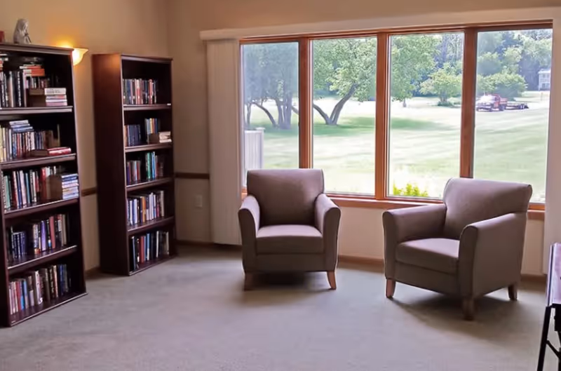 A cozy sitting area with two upholstered armchairs positioned near a large window overlooking a green outdoor landscape. To the left, there are two tall wooden bookshelves filled with books, and a floor lamp providing warm lighting.