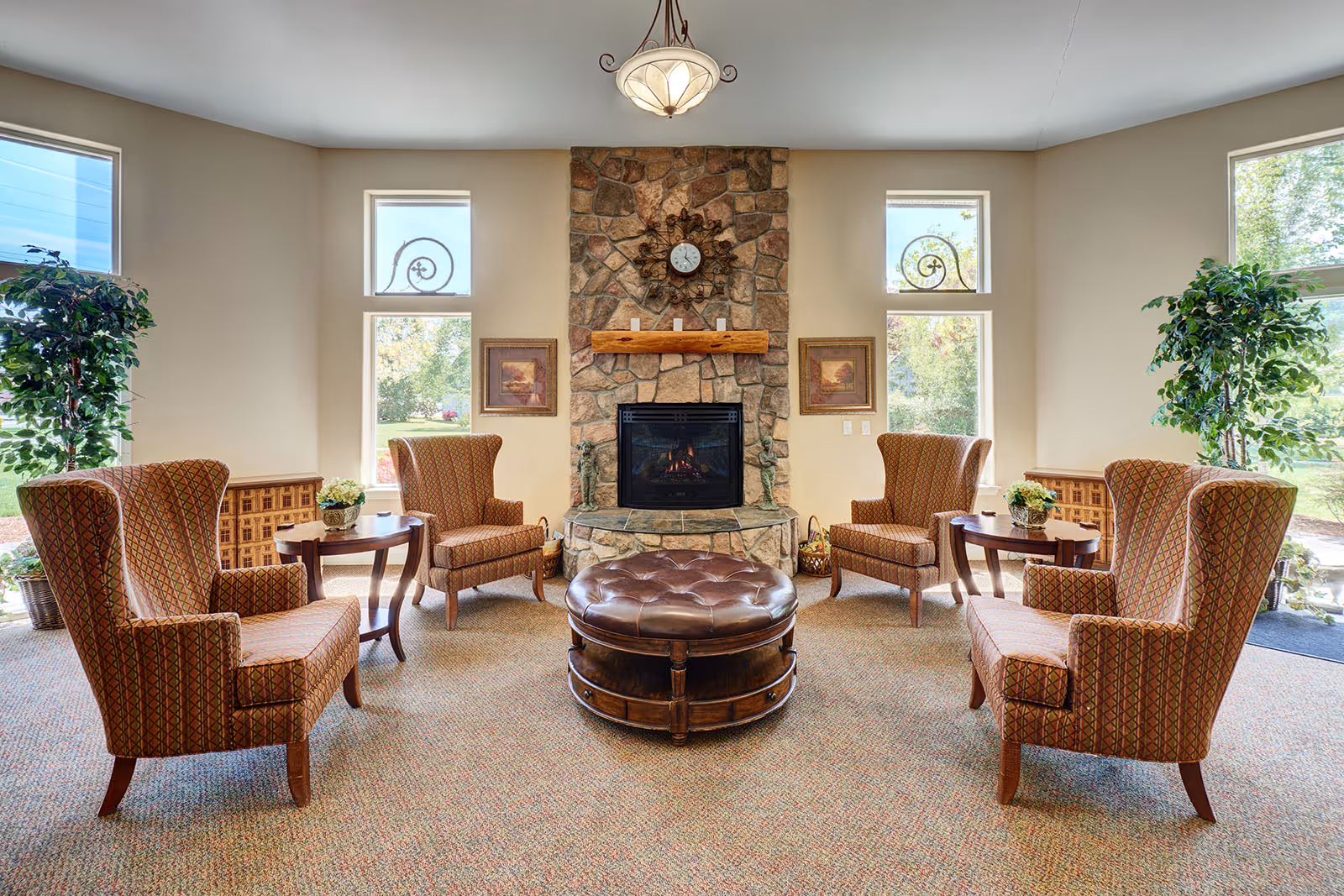 A cozy living room area with four patterned armchairs arranged around a round leather ottoman. The room features a stone fireplace with a wooden mantel, a clock above it, and two framed pictures on either side. Large windows let in natural light and there are two tall potted plants near the windows.