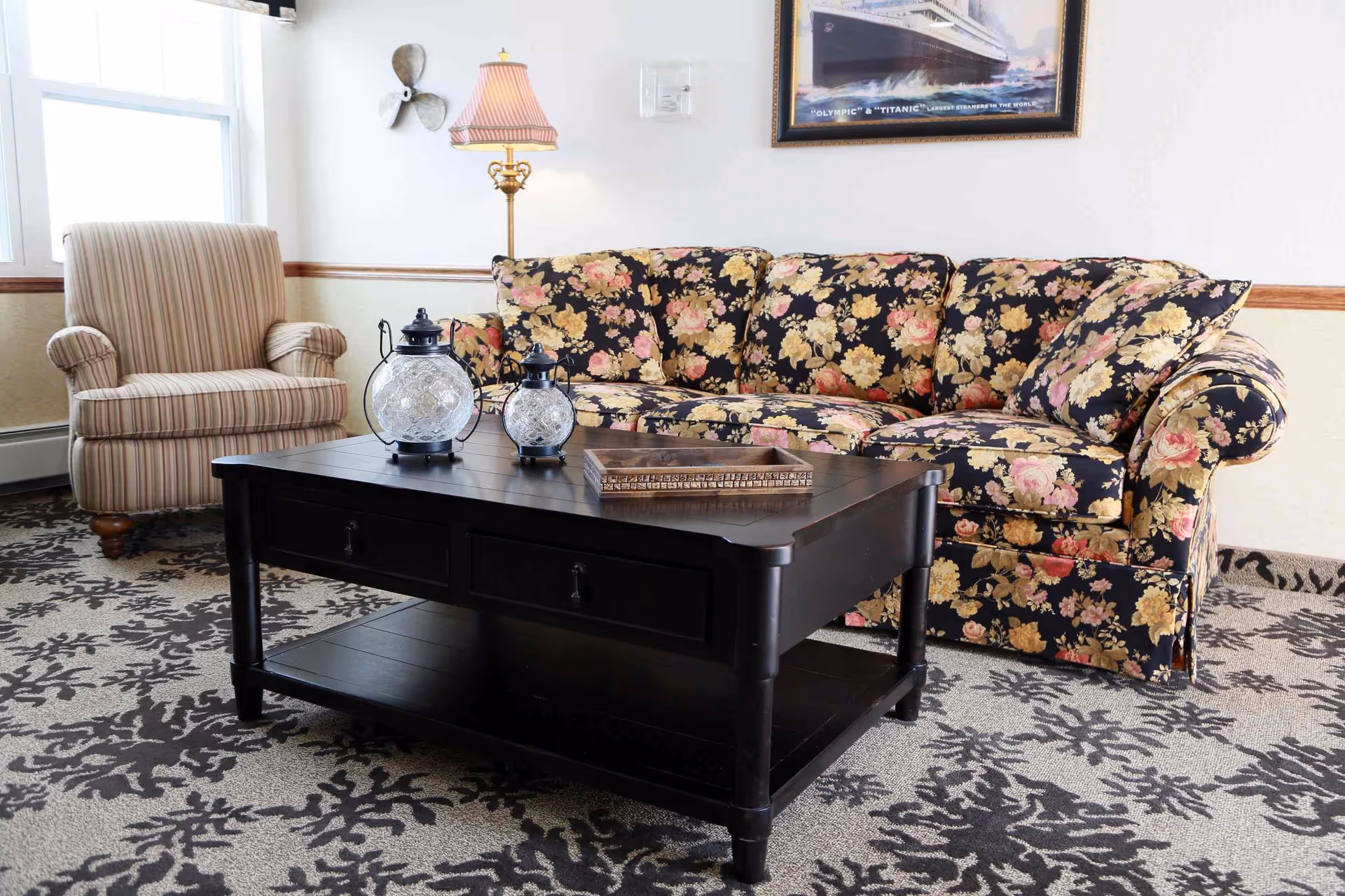 A cozy living room area featuring a floral patterned sofa and a striped armchair. In front of the sofa is a dark wooden coffee table with two decorative glass lanterns and a rectangular tray. The room has a patterned carpet and a framed picture of a ship on the wall above the sofa. A floor lamp with a pleated shade stands next to the sofa.