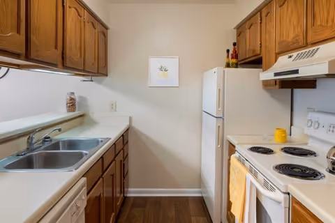 A compact kitchen with wooden cabinets, a double stainless steel sink, a white refrigerator, an electric stove with four burners, and a white range hood. The kitchen has a light-colored countertop and a small framed picture on the wall. There are a few decorative items on top of the refrigerator and a yellow towel hanging on the oven door.