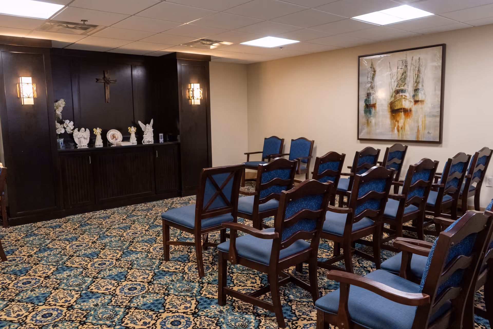 A small chapel or meditation room with rows of wooden chairs with blue cushions facing a dark wooden altar adorned with religious statues and a cross on the wall. The room has patterned carpet flooring and a painting of boats on the wall.