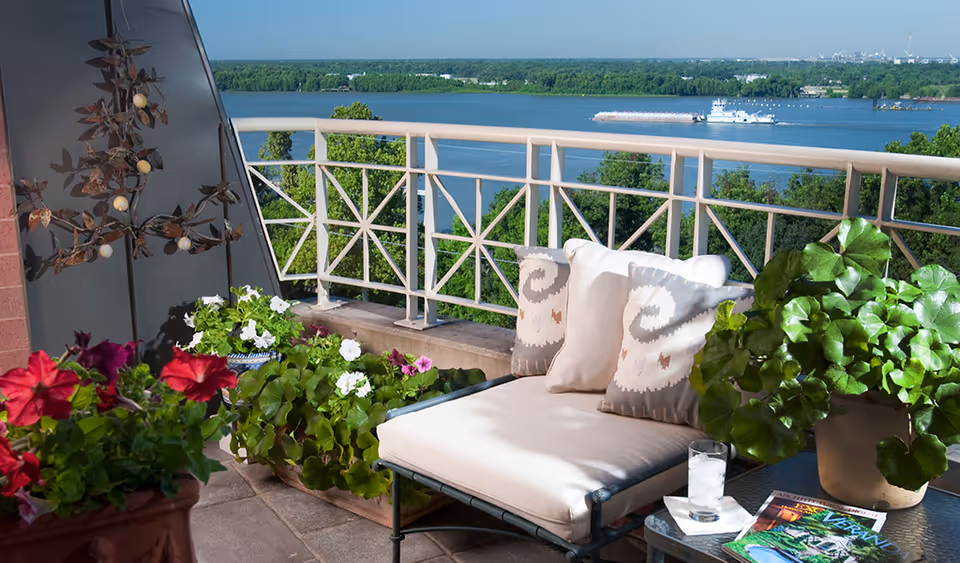 A balcony terrace with a cushioned lounge chair, potted flowers and plants, and a river view with a boat in the distance.