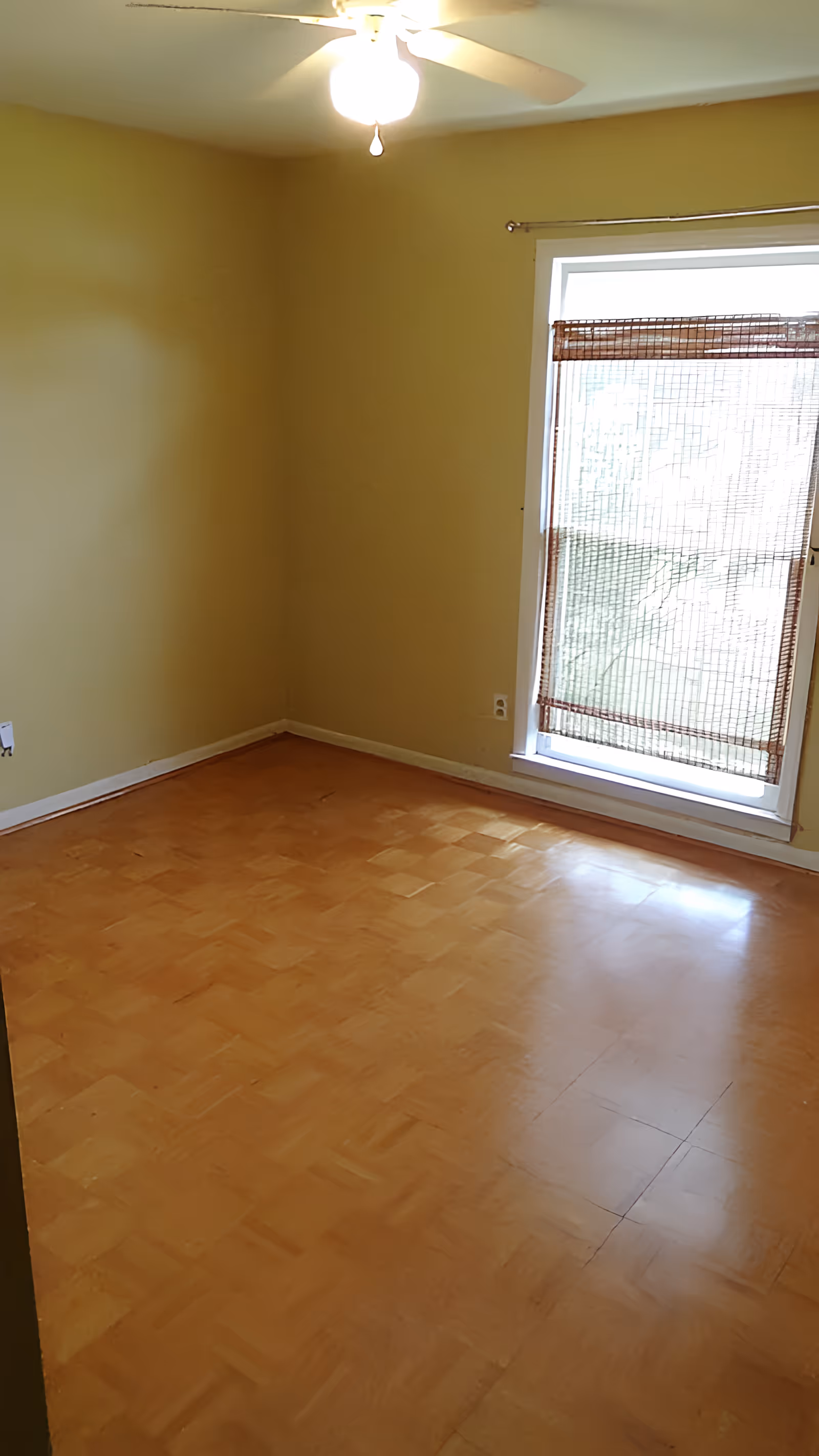 Empty room with light brown parquet flooring, pale yellow walls, a ceiling fan with light, and a window covered by a bamboo blind.