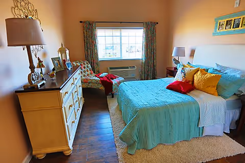 A cozy bedroom with a bed covered in a turquoise quilt and colorful pillows, a beige dresser with a lamp and decorative items, a patterned armchair with a red blanket, a window with floral curtains, and wooden flooring with a light-colored rug.