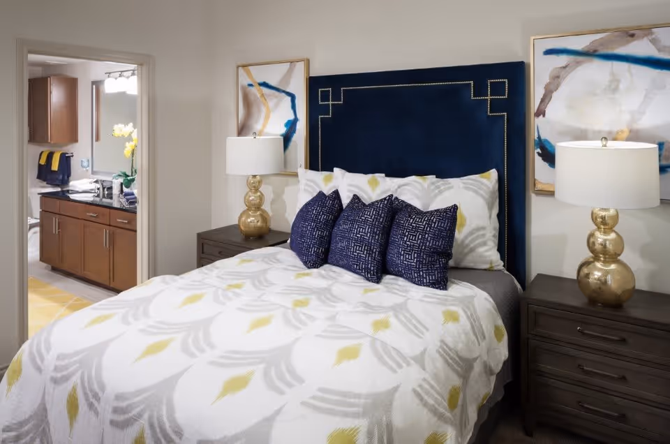 A neatly made bed with a dark blue upholstered headboard and white bedding featuring a gray and yellow leaf pattern. There are three dark blue decorative pillows on the bed. On each side of the bed are dark wooden nightstands with gold lamps. Two abstract paintings hang on the wall above the nightstands. In the background, an open doorway reveals a bathroom with wooden cabinets, a sink, and a mirror.