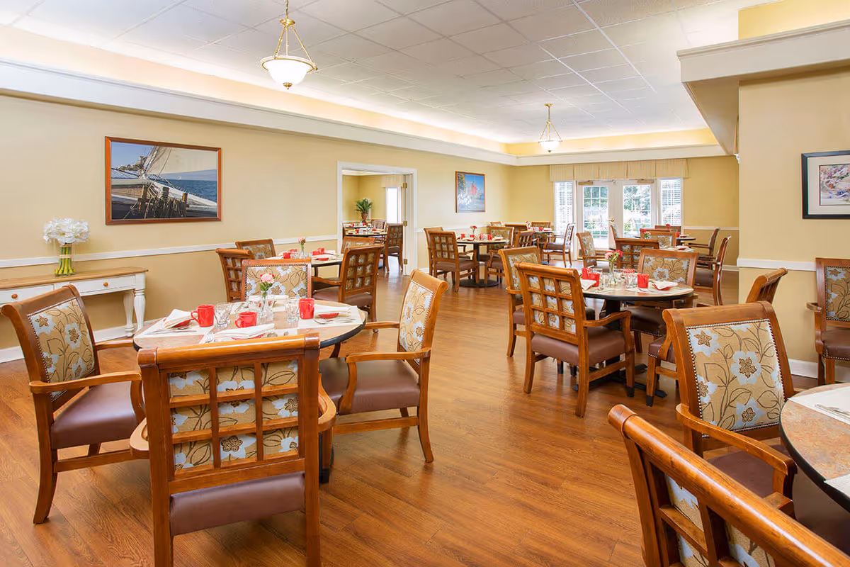 A bright dining room with multiple round tables set with red cups, napkins, and silverware. The room features wooden chairs with floral patterned upholstery, wood flooring, beige walls, and framed artwork. Large windows allow natural light to fill the space.