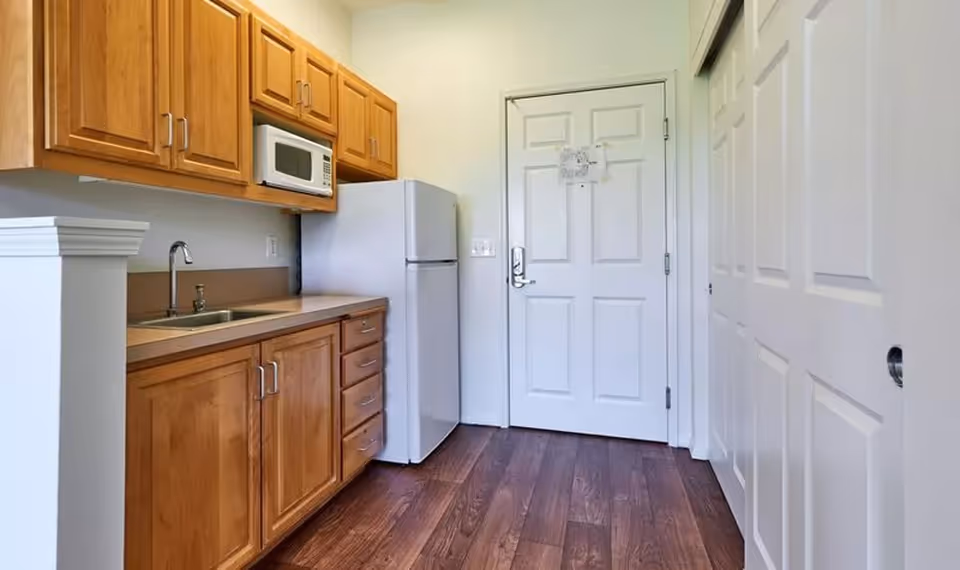 A small kitchen area with wooden cabinets, a countertop with a sink, a microwave mounted above the counter, and a white refrigerator. The floor is dark wood, and there is a white door and white closet doors on the right side.