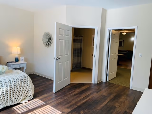 A bedroom with a bed covered in a chunky knit blanket, a nightstand with a lamp and picture frame, and two open doors. One door leads to a bathroom with a shower curtain visible, and the other door opens to a hallway or another room with carpeted flooring.
