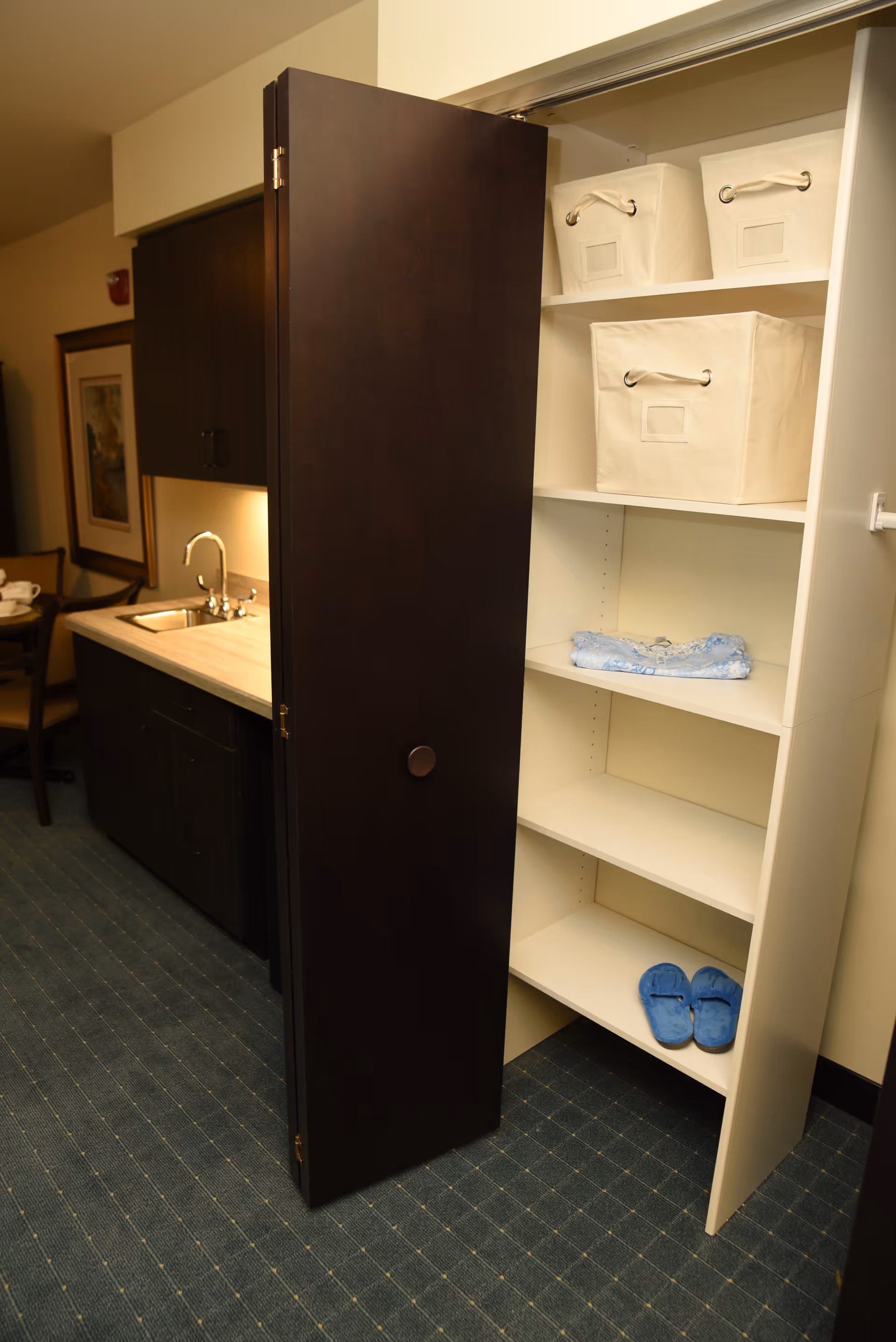 An open dark wood folding door reveals white built-in shelves with fabric bins and blue slippers beside a small kitchenette with a sink.