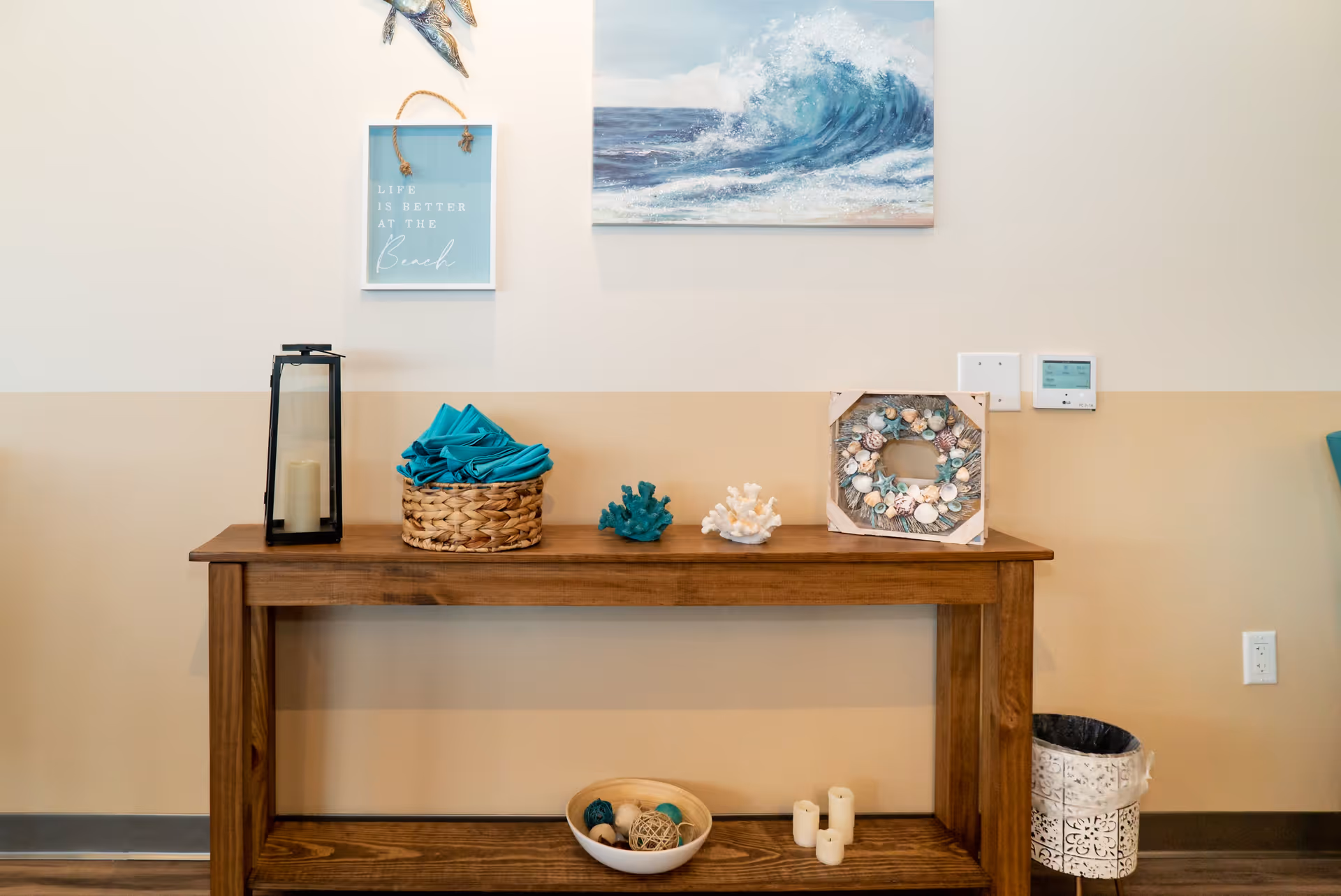 A wooden console table against a beige and white wall with beach-themed decorations including a basket with teal napkins, coral ornaments, a seashell wreath, a lantern with a candle, and a bowl with decorative balls. Above the table are two wall hangings: one with a wave painting and another with the text 'Life is better at the Beach'. A small trash can is placed on the floor to the right of the table.
