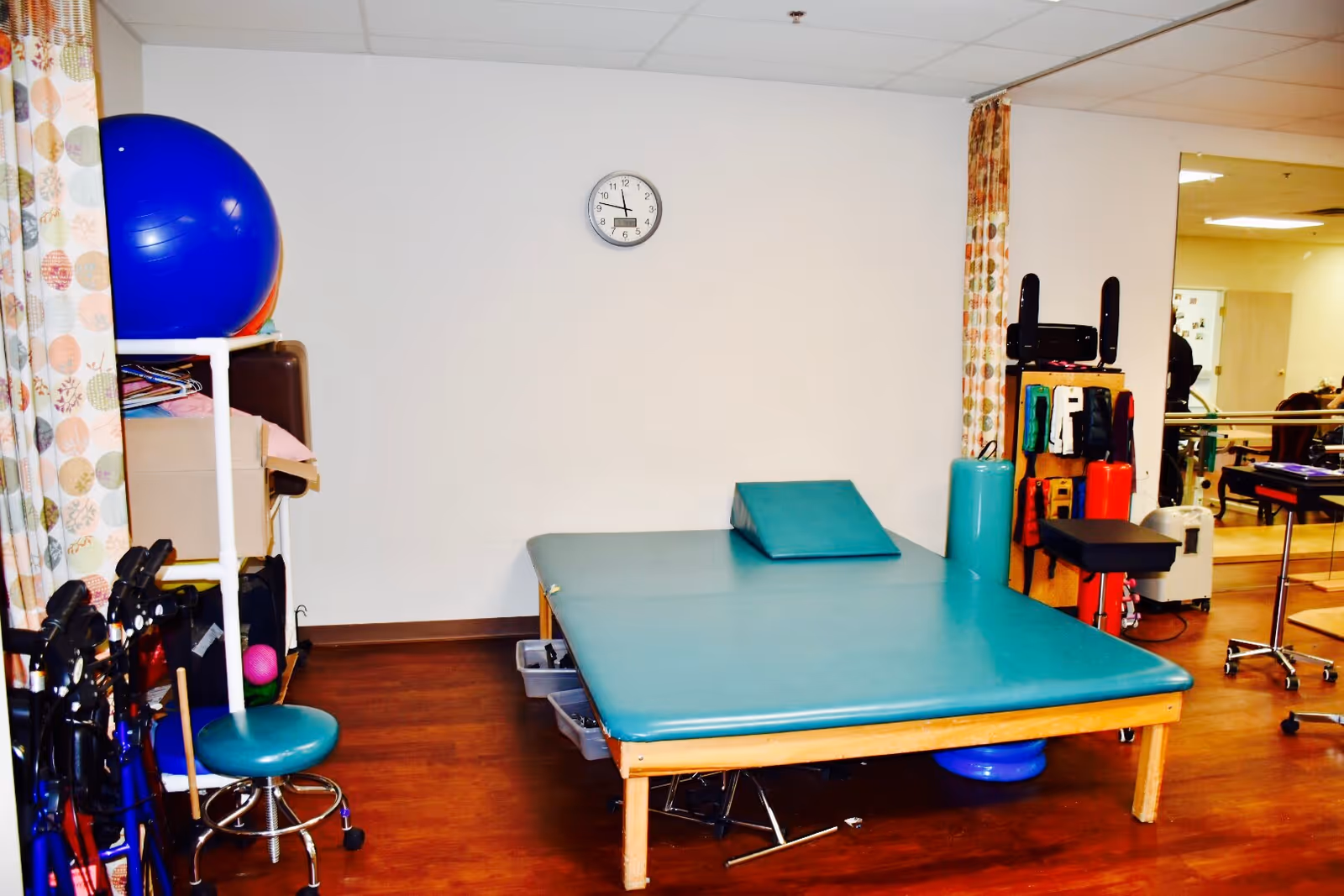 A physical therapy room with a large padded treatment table, a blue exercise ball, various therapy equipment, and a clock on the wall showing 4:45. The room has wood flooring and light-colored walls with patterned curtains.