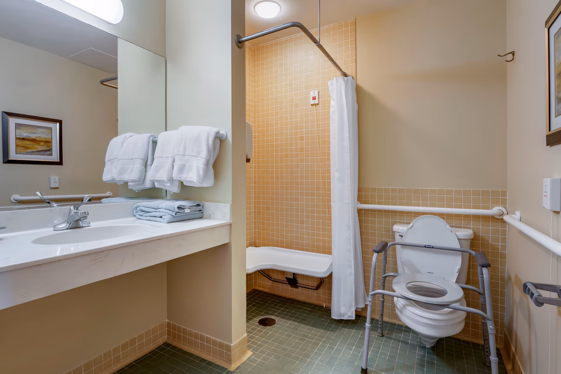 Accessible bathroom with a wall-mounted sink and faucet, a large mirror above the sink with white towels hanging on a rack, a shower area with a white curtain and a fold-down shower seat, and a toilet equipped with safety rails and a raised seat. The walls have beige tiles and the floor has green tiles. A framed picture is hung on the wall.