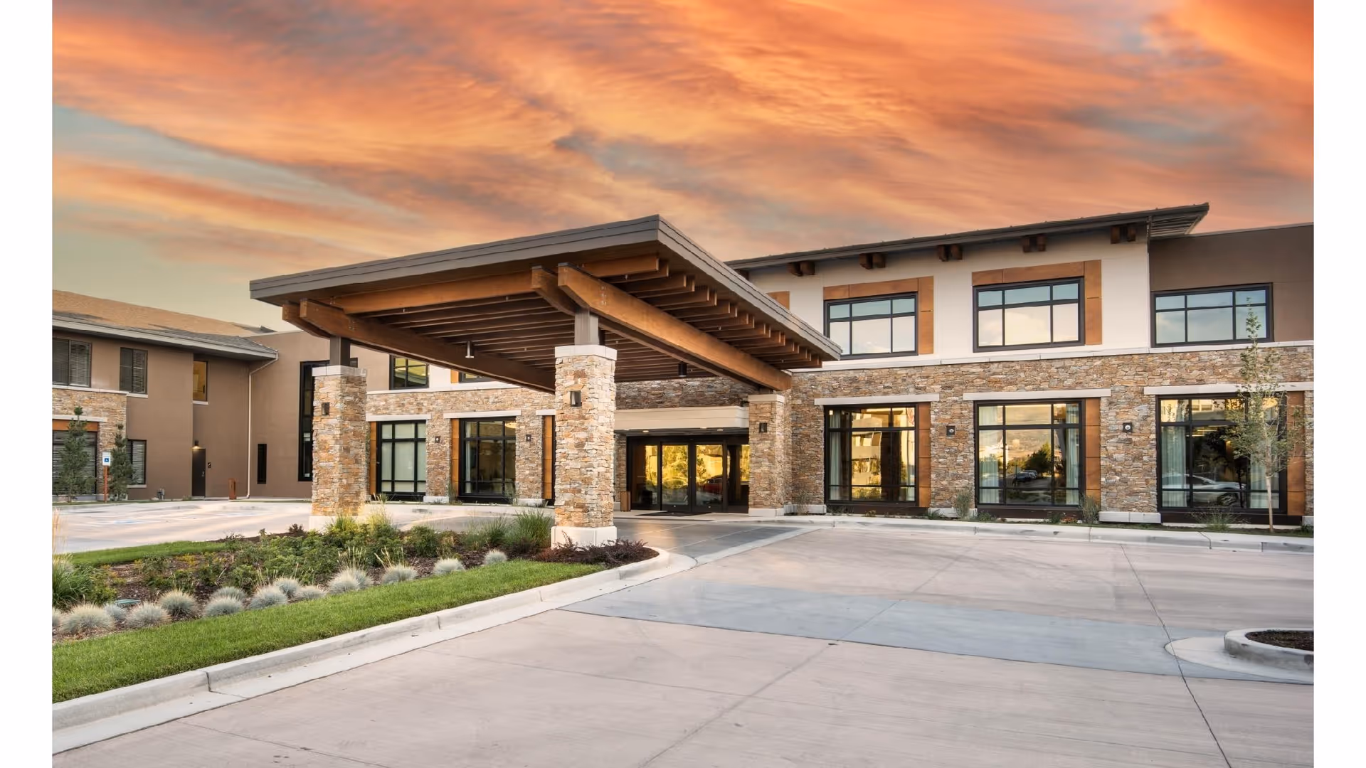 Front entrance of a modern senior living building with a covered porte-cochere, stone facade, large windows, landscaping, and a colorful sunset sky.