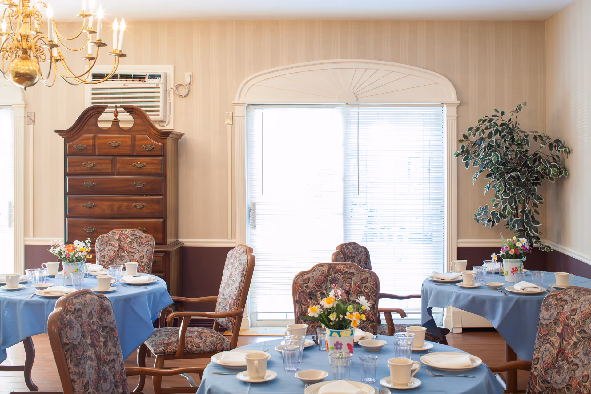 A dining room with round tables covered in blue tablecloths, set with white plates, cups, and glasses. Each table has a small floral centerpiece. The room features floral upholstered chairs, a large wooden chest of drawers, an air conditioning unit, a chandelier, and a large window with blinds. There is also a large potted plant in the corner.