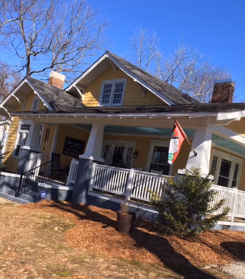 Yellow bungalow-style house with a wide front porch, white railings, a festive flag, and bare trees under a clear blue sky.