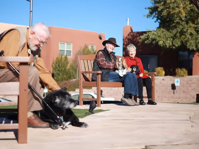 Two elderly people sitting on a bench outside a building, each holding a small dog. Another elderly man is sitting on a separate bench nearby, petting a black dog lying on the ground. The setting is sunny with a clear sky, and the building in the background has a southwestern architectural style.