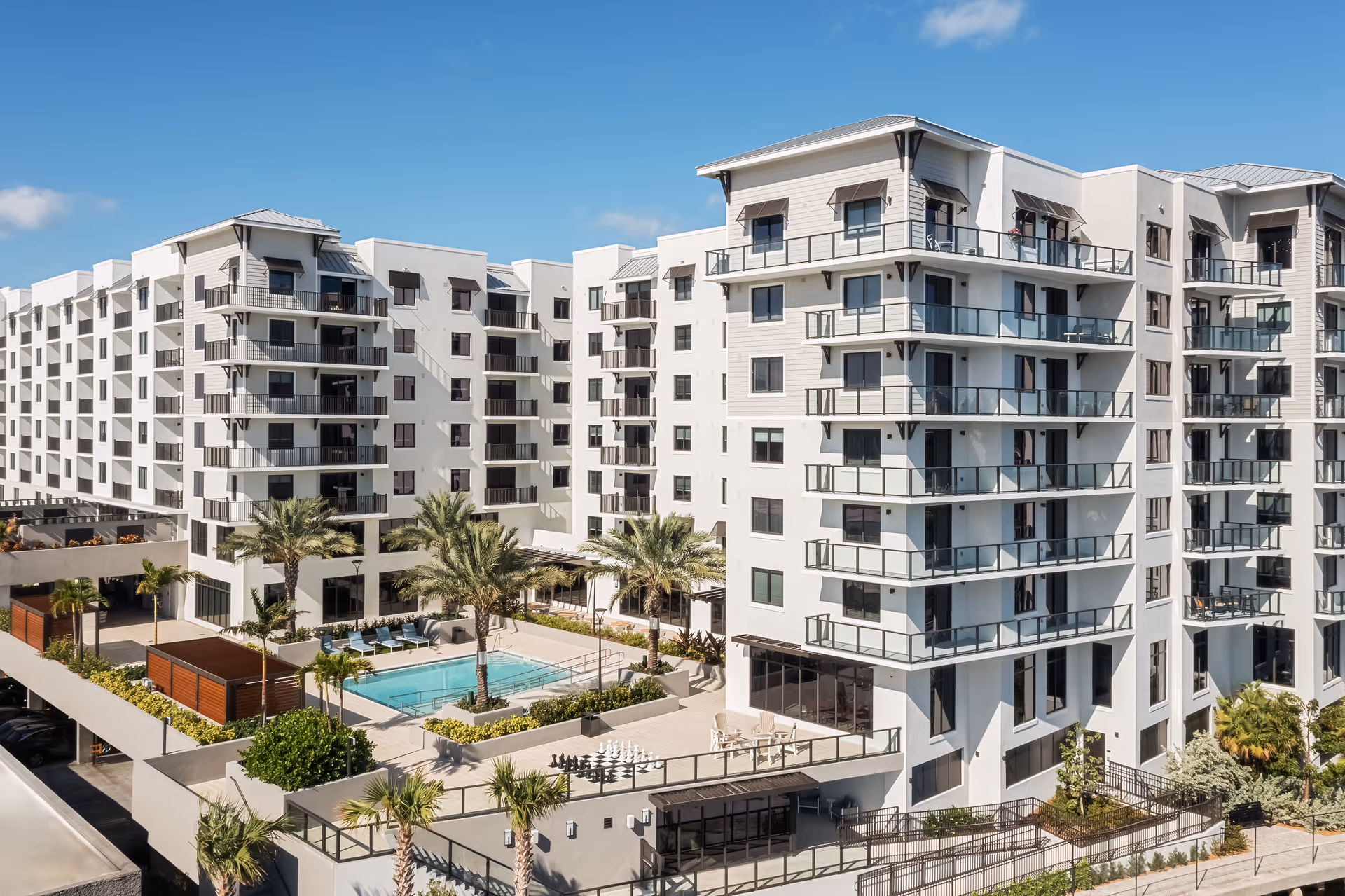 Modern multi-story residential building complex with balconies, a swimming pool, palm trees, lounge chairs, and a large outdoor chess set under a clear blue sky.