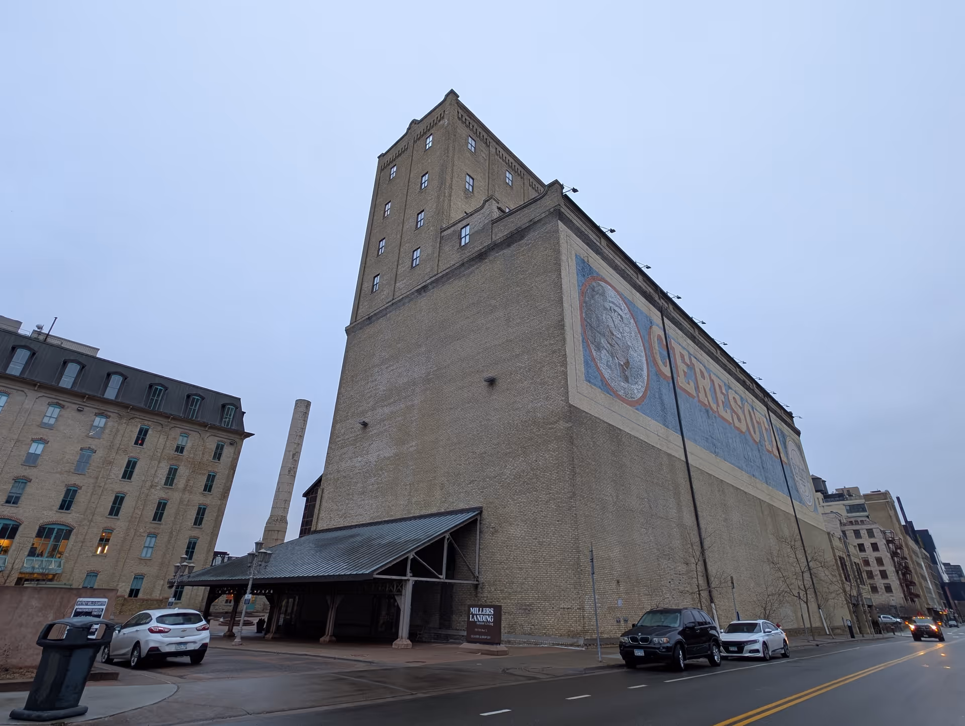 Tall historic brick building featuring a large painted advertisement on its side, a covered entrance, and parked cars along a city street.