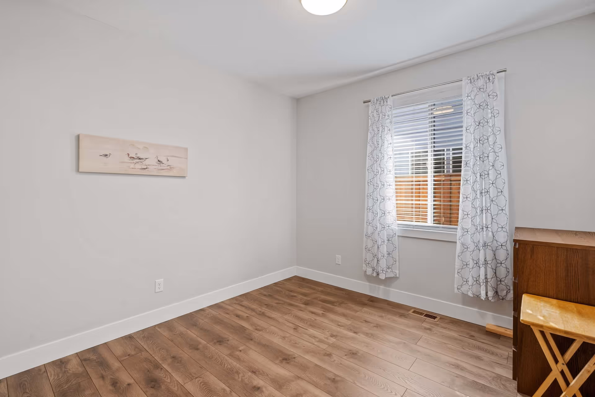 A small, empty room with light gray walls, wood flooring, a window with white patterned curtains, a wooden cabinet, and a small wooden stool. A painting of birds is hung on the wall.