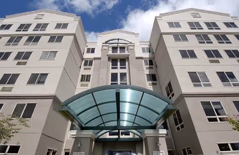 Front exterior of a multi-story building with a curved glass entrance canopy under a partly cloudy sky.